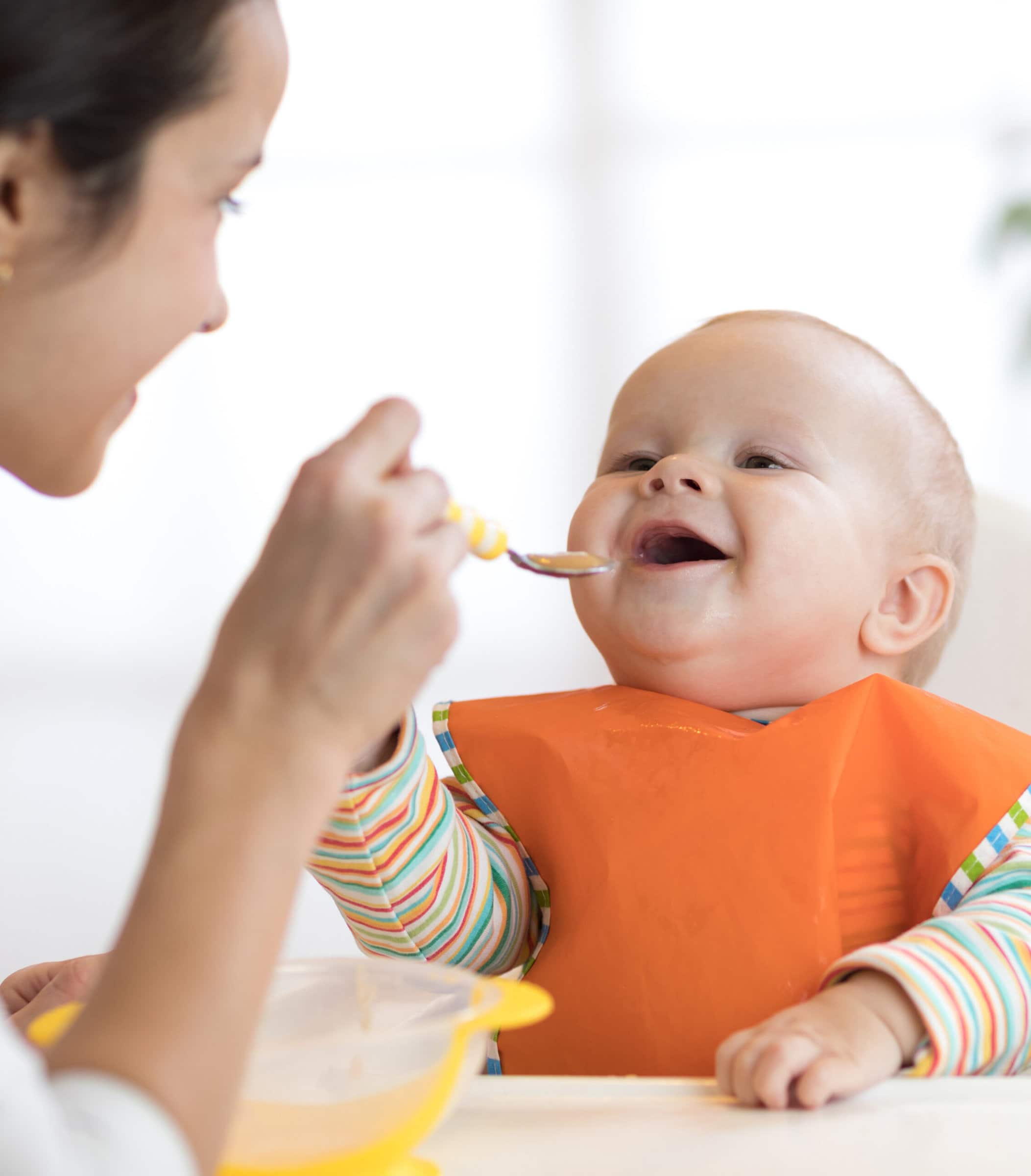 baby in high chair and orange bib being fed by their mother