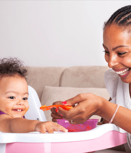 Smiling mother feeding baby in high chair with orange spoon