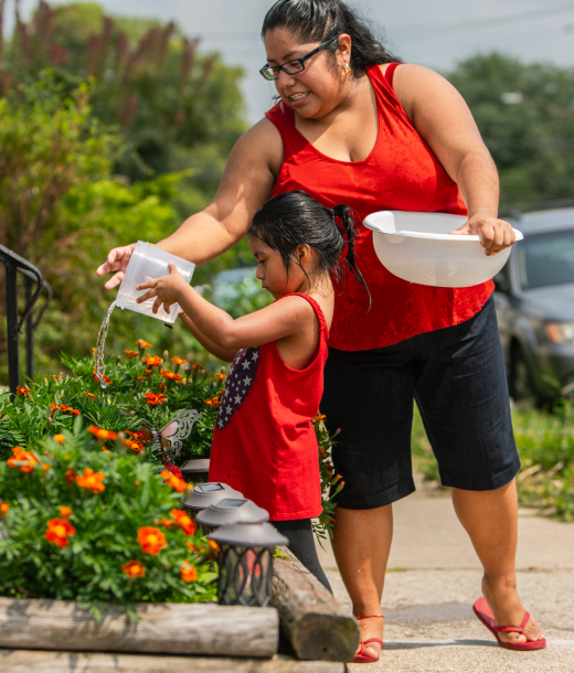 Daughter and mother in red dresses watering plants in front yard on city street