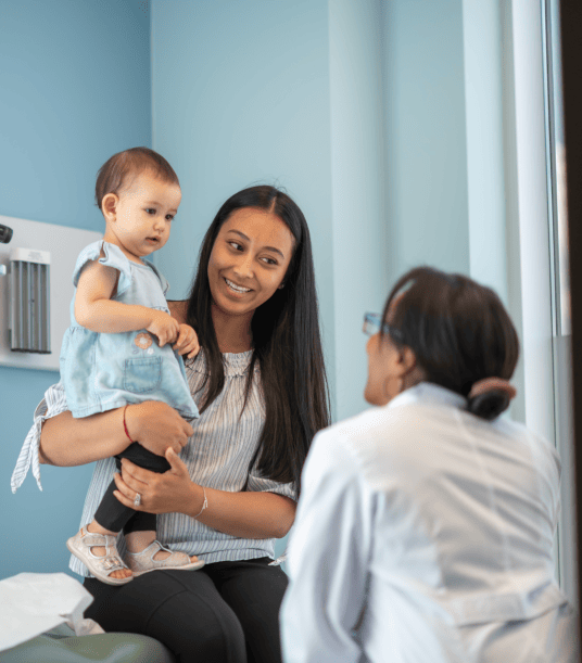 Mother with long straight black hair holding baby on exam table in doctor's office as doctor looks on
